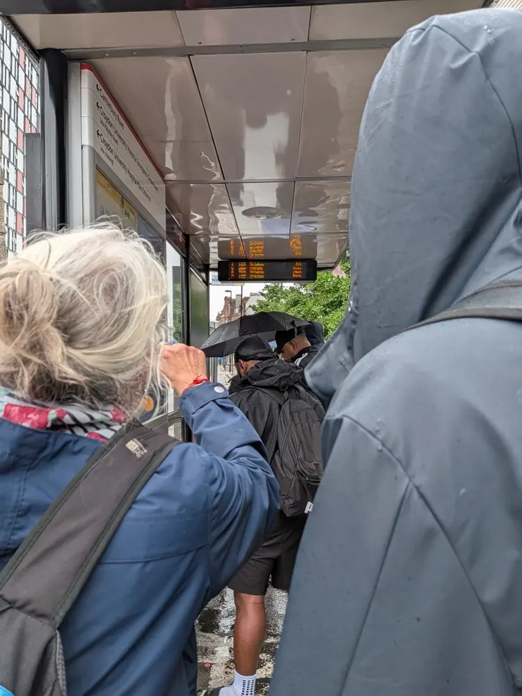 People queuing at a rainy London bus stop beneath a digital board showing bus arrival times.